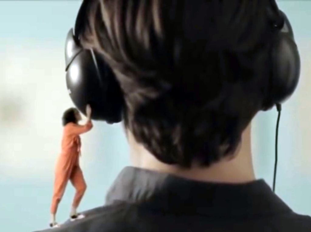 A tiny woman stands on a man's shoulder, trying to listen to his headphones.