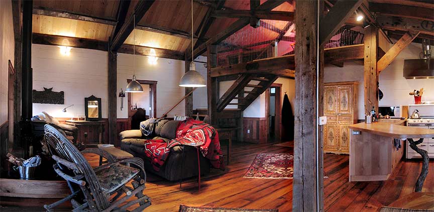 Messy, decorative artist's loft with hardwood floors, a carved armoire, blankets thrown over a plush couch, and dark wood support beams against white brick walls.