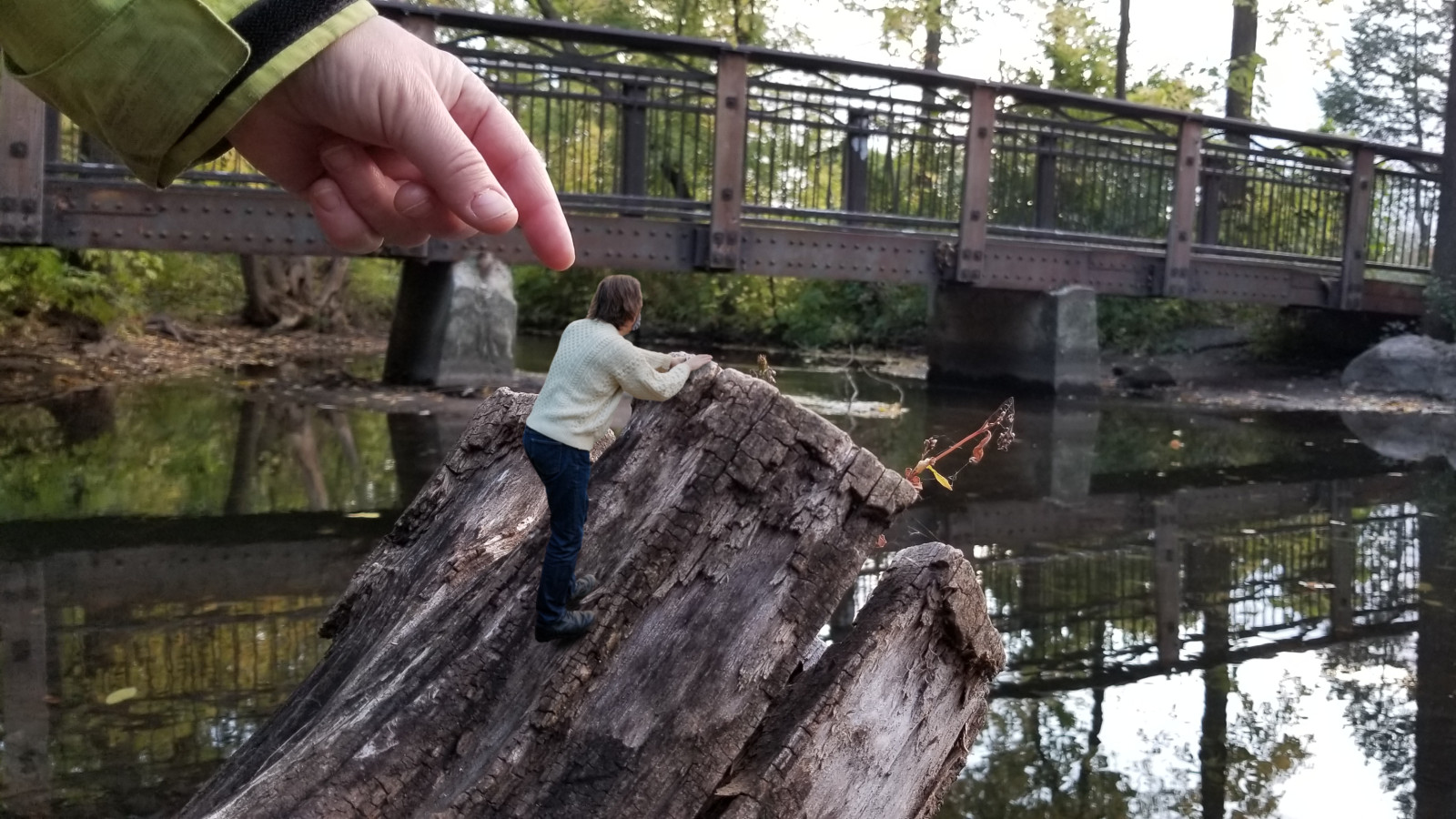 A tiny man in a sweater climbs a stump by a creek. A gigantic woman's hand reaches down to pick him up.