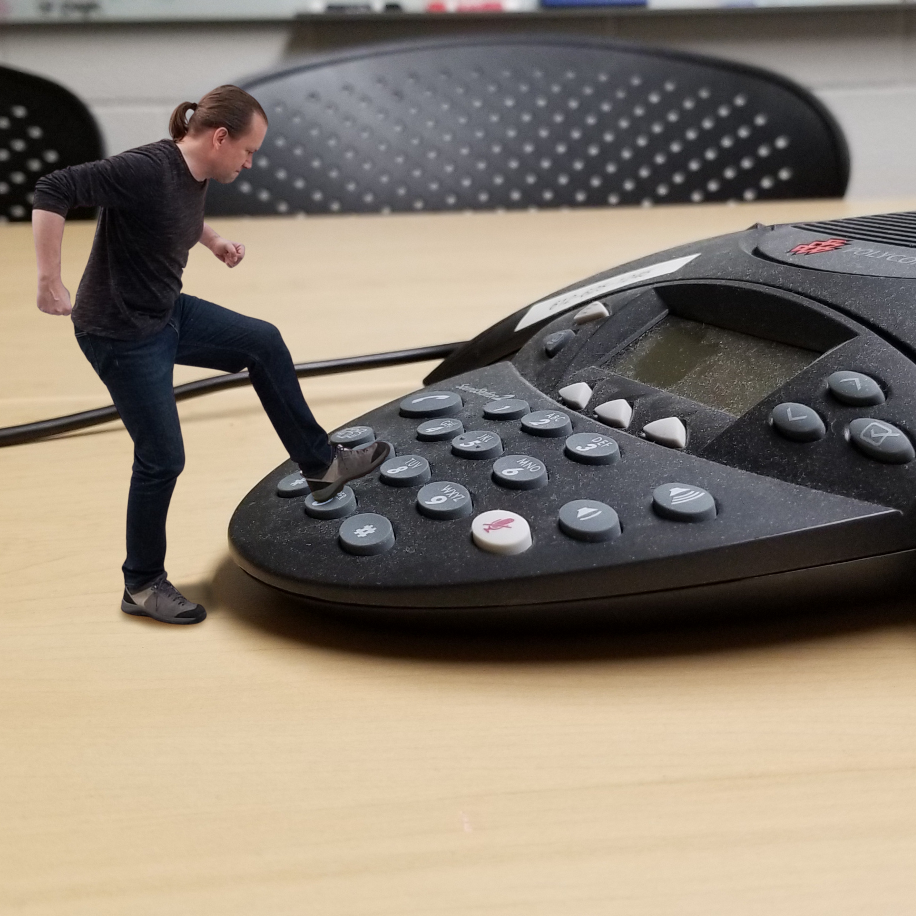 A tiny man in dark clothes stomps on the keypad of an office conference phone.